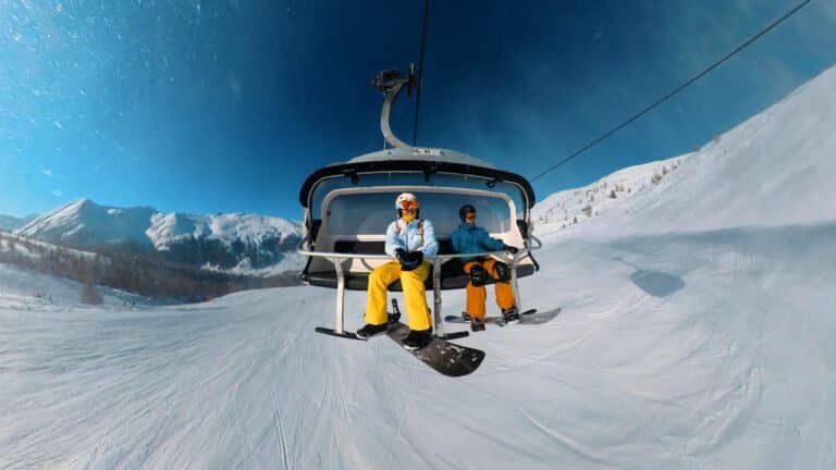 Two snowboarders ascending a snowy slope on a ski lift in Livigno, Italy under clear blue skies.